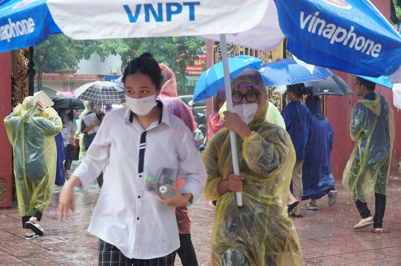 Một nữ thanh niên tình nguyện mang áo mưa tiện lợi, tay cầm chiếc ô to để che mưa cho thí sinh tại điểm thi Trường THPT Huỳnh Thúc Kháng (TP Vinh, Nghệ An).