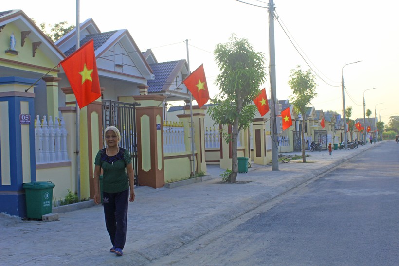 Bà Nguyễn Thị Hoạt sải bước trước khu nhà xây mới, rực sắc đỏ cờ Tổ quốc. 